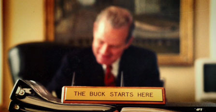 Baker at desk with desk plaque that reads, the Buck Starts Here