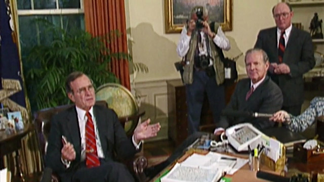 President Bush seated in chair with Baker and reporters gathered around.